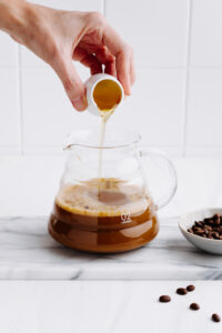 A glass pot of coffee with milk with coffee beans next to it on a white backdrop and a hand pouring maple syrup in it
