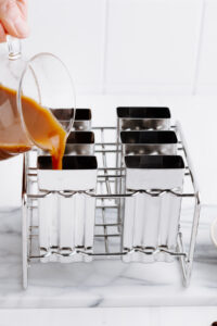 Stainless steel popsicle molds on a white backdrop with a glass pot pouring coffee with milk in a mold