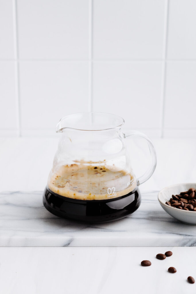 A glass pot of dark coffee with coffee beans next to it on a white backdrop with white tiles