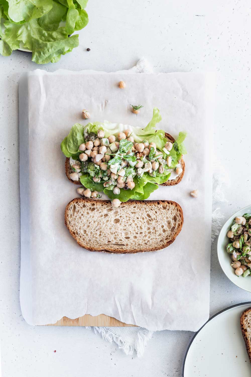 Chickpea Salad Sandwich on a piece of bread with a piece of lettuce next to a slice of bread on white parchement paper