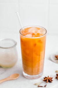 Bright orange colored Thai tea in a tall glass with ice cubes and a glass straw on a white backdrop next to a small wooden spoon, spices and a small jar with milk.