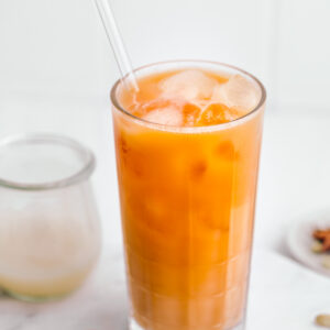 Bright orange colored Thai tea in a tall glass with ice cubes and a glass straw on a white backdrop next to a small wooden spoon, spices and a small jar with milk.