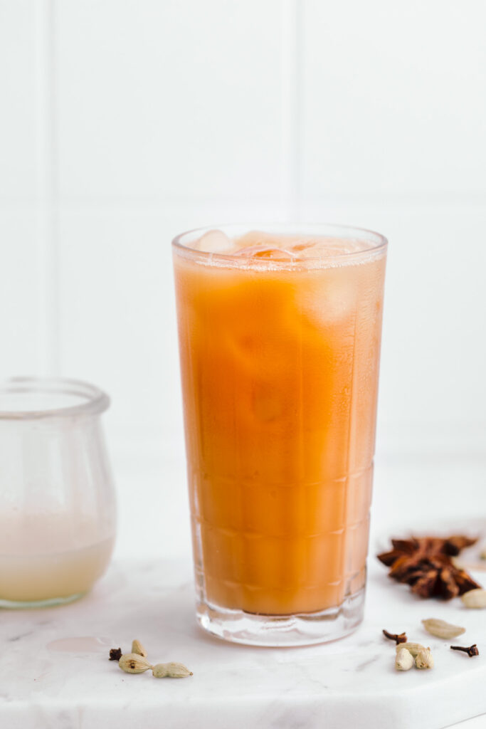 Bright orange colored Thai tea on a white marble backdrop in a tall glass with ice cubes next to star anise, cardamom and cloves and a small jar with milk.