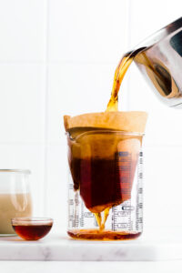 A glass measuring cup on a white backdrop with a brown coffee filter in it and a stainless steel pan pouring in black tea.