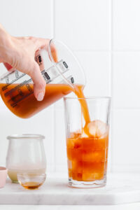 A tall glass with ice cubes on a white marble cutting board with a hand pouring in orange Thai tea in the glass with a glass measuring cup.