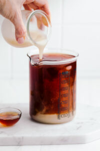 A glass measuring cup on a white marble cutting board with a layer of milk on the bottom and the rest filled with black tea and a hand pouring in milk from a small glass cup.
