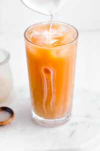 Bright orange colored Thai tea in a tall glass with ice cubes on a white backdrop and the tip of a small white bowl pouring in milk on top of the tea.