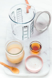 A white marble cutting board with a glass measuring cup, a tea filter, a small glass bowl with syrup, a small glass jar with condensed milk, a small pink bowl with milk and a wooden spoon.
