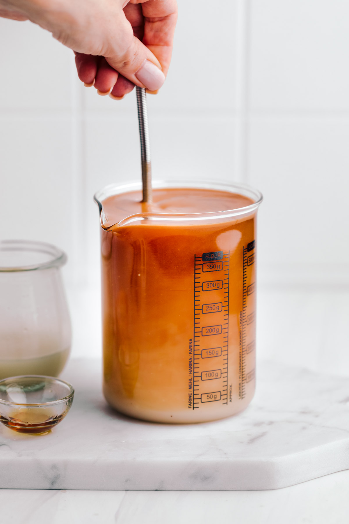 A glass measuring cup filled with light orange Thai tea with a hand whisking the tea on a white marble backdrop with a small glass bowl next to it.