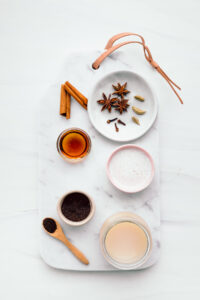 A white marble cutting board with cinnamon, cloves, star anise, cardamom, milk, syrup, black tea and sweetened condensed milk in small bowls and on a small white plate.