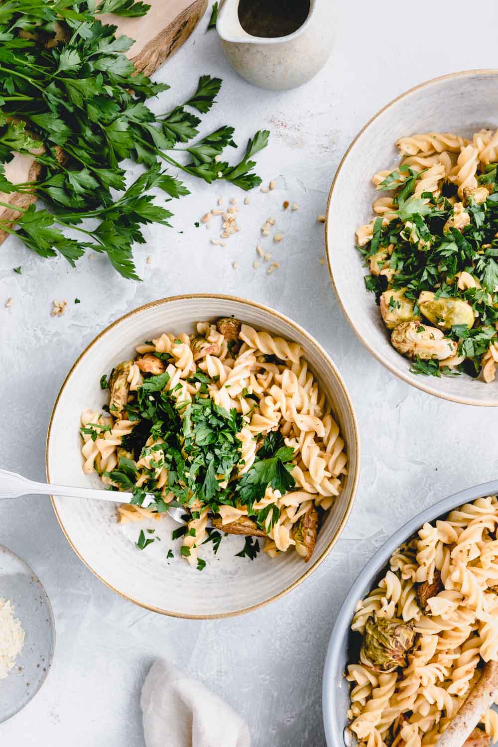 Two bowls with Brussels sprout pasta and chopped parsley on a white backdrop and a small edge of a pasta pan in the corner and fresh parsley on a wooden cutting board in the other corner