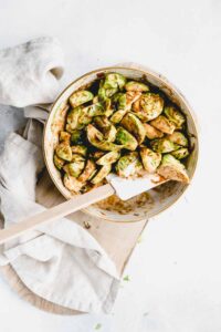 Brussels sprouts cut and coated in a bowl with a wooden spatula on a white backdrop, a light wooden cutting board and a light napkin
