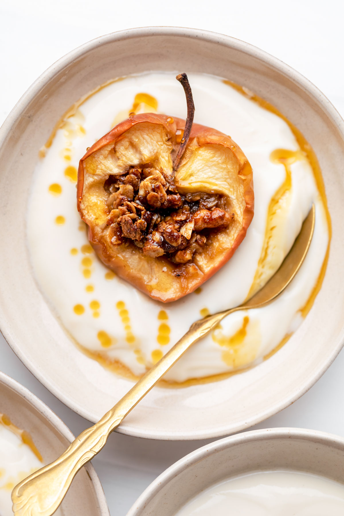 baked apple in a bowl with yogurt drizzled with maple syrup and a gold teaspoon