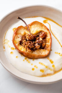 baked apple with filling in a beige bowl with soy yogurt on a white backdrop drizzled with maple syrup