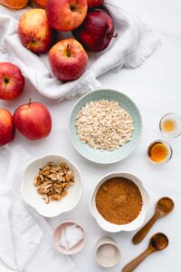 ingredients for baked apples in small bowls and wooden spoons on a white backdrop with a white napkin