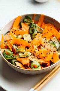 Cucumber and carrot salad sprinkled with white and black sesame seeds in a light brown bowl on a white plate with two wooden chopsticks
