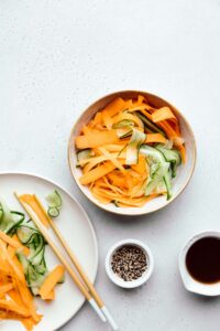 Cucumber and carrot ribbons in a light brown bowl on a light grey backdrop with a plate with cucumber and carrot ribbons and a small bowl with sesame seeds and soy sauce