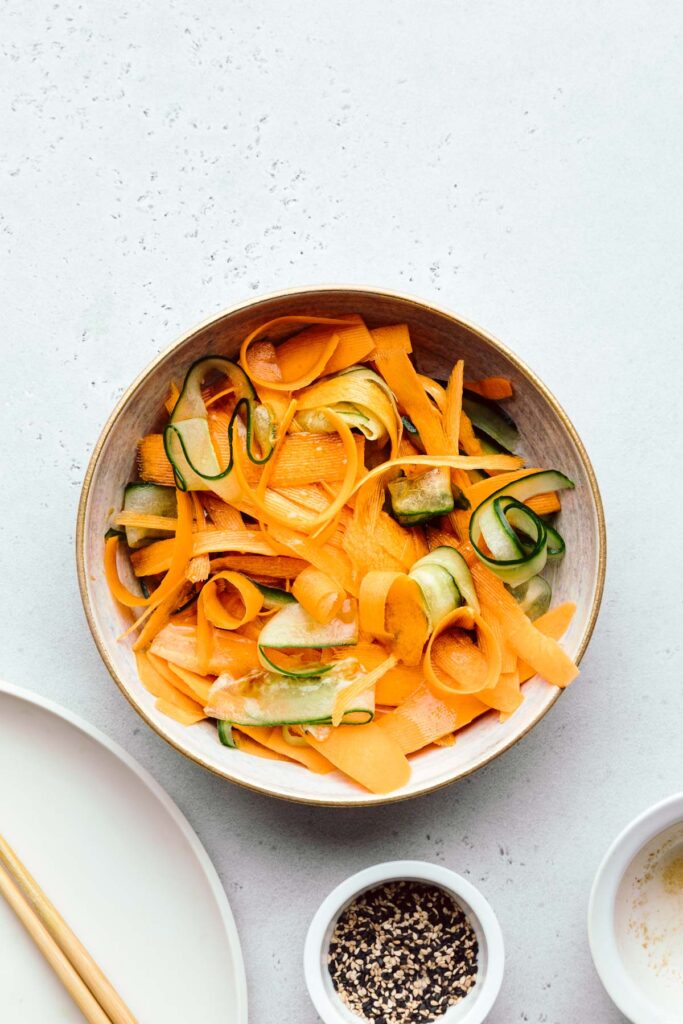 Cucumber and carrot ribbons in a light brown bowl on a light grey backdrop with the dressing on it and a small bowl with sesame seeds