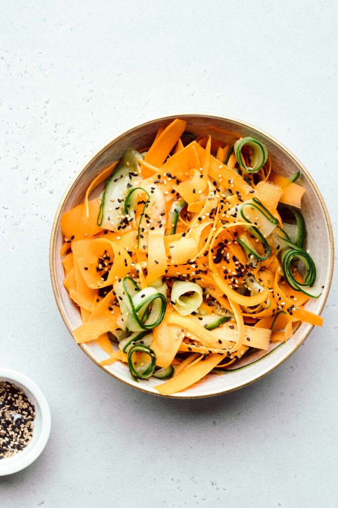 Cucumber and carrot salad sprinkled with white and black sesame seeds in a light brown bowl on a light backdrop with a small white bowl with sesame seeds next to it