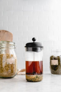 a french press with loose leaf tea and two jars of tea herbs next to it in front of a white backdrop