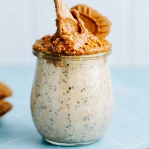 Dipping a wooden spoon in a small glass jar with overnights oats and creamy Lotus Biscoff spread and cookie as a topping.