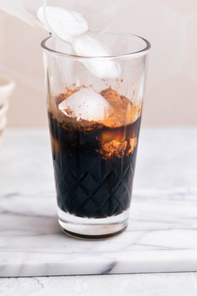 A glass cocktail shaker with espresso and ice cubes falling into the cocktail shaker on a white marble backdrop.