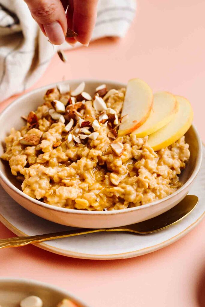 Brown Sugar Cinnamon Oatmeal topped with sliced apples and chopped almonds being sprinkled over it.