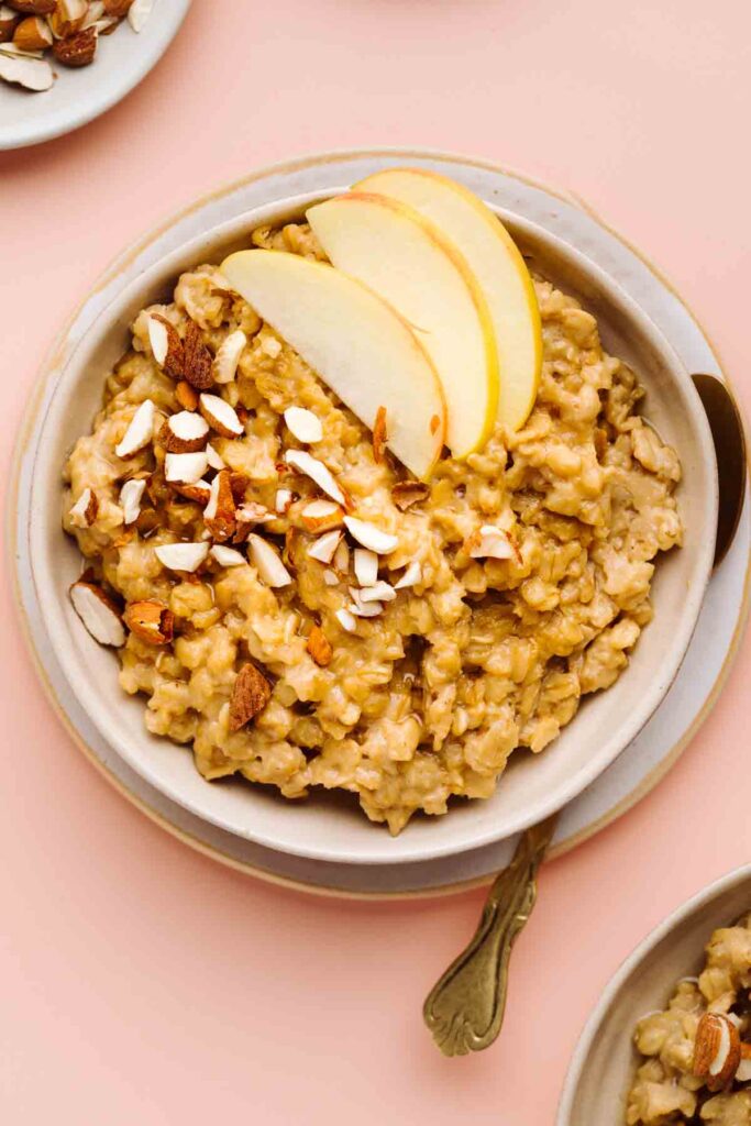 Brown Sugar Cinnamon Oatmeal topped with sliced apples and chopped almonds in a bowl on a plate with a gold colored spoon next to it on a peach colored backdrop.