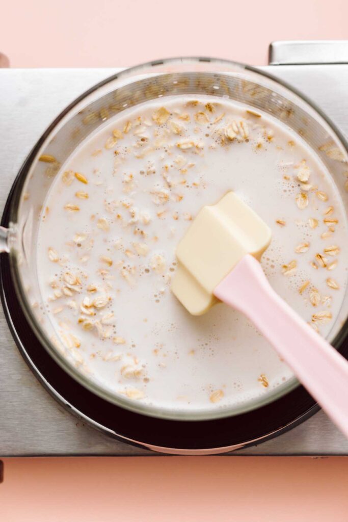 A transparent pan on a stove top with oats, milk and a spatula in it.