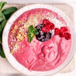 Raspberry Smoothie Bowl in a white bowl topped with mint leaves, nuts, jam and coconut flakes next to fresh mint leaves on a wooden backdrop