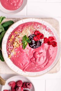 Raspberry Smoothie Bowl in a white bowl topped with mint leaves, nuts, jam and coconut flakes next to fresh mint leaves on a wooden backdrop