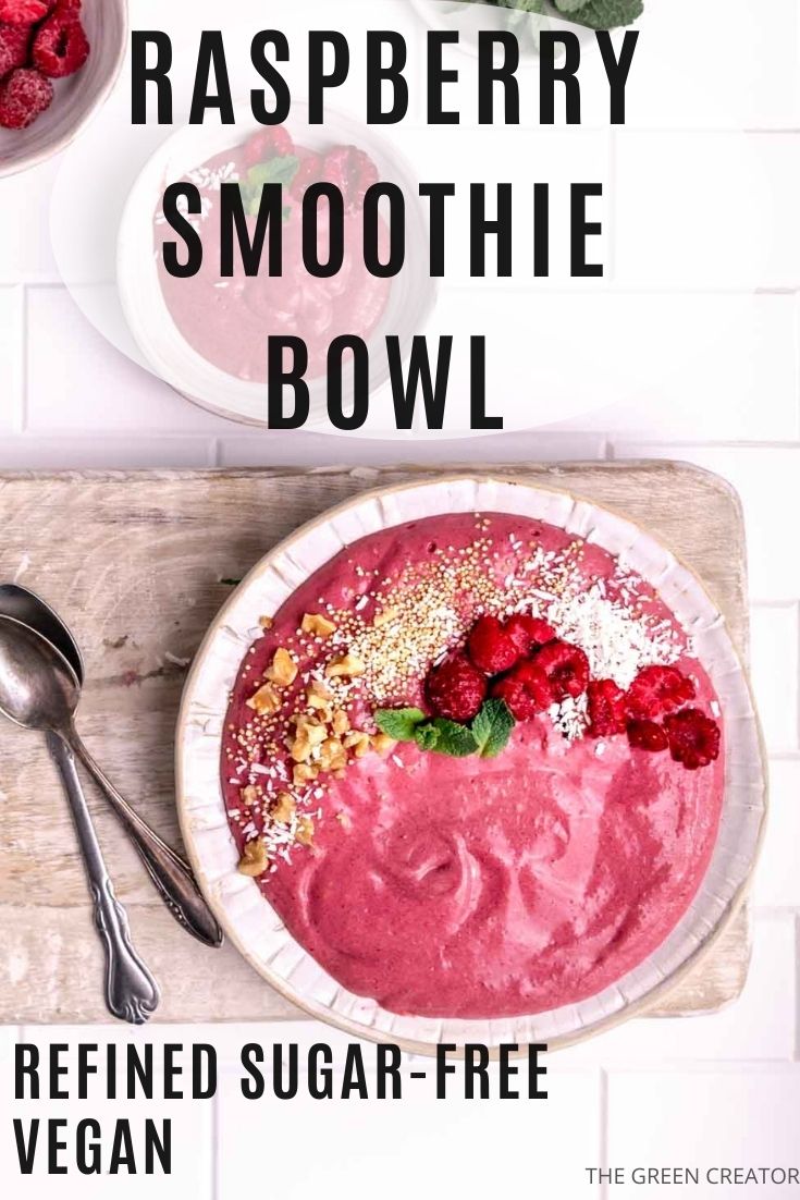 Raspberry Smoothie Bowl in a white bowl topped with mint leaves, nuts, jam and coconut flakes next to two teaspoons on a wooden backdrop and a small bowl with raspberry smoothie