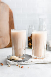 two tall glasses with banana bread milk on a white backdrop with a white napkin and a small light blue plate with a few slices of banana and nuts in the background