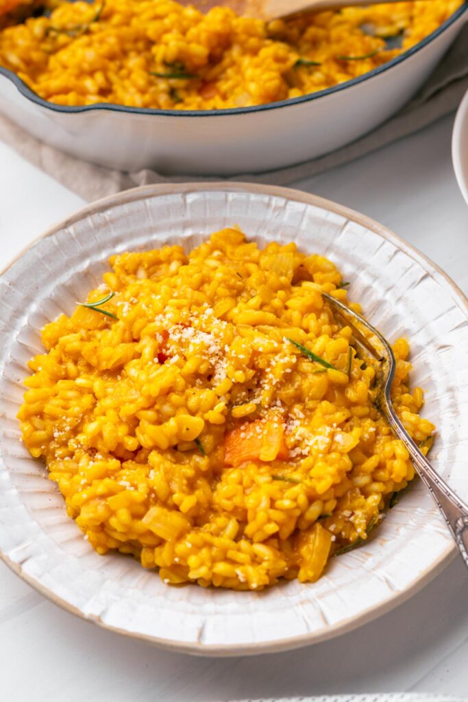 Creamy vegan pumpkin risotto in a cream-colored bowl with a fork on a white backdrop with a cast iron pan with risotto in the back