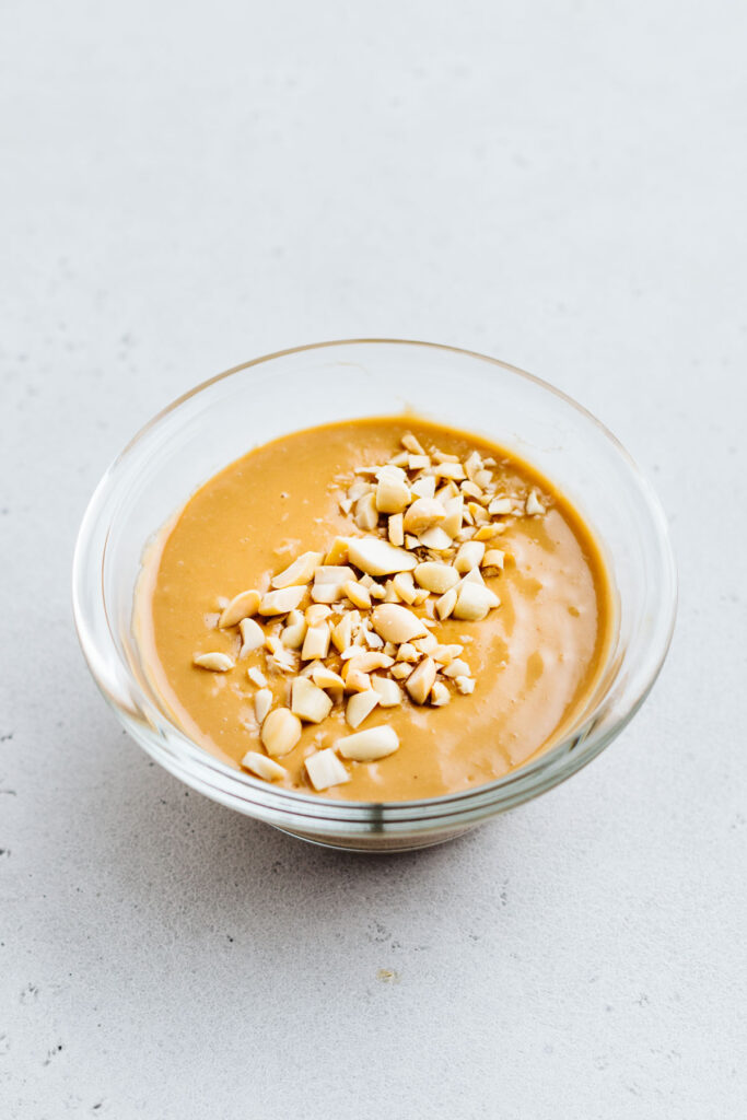 A small glass bowl on a grey backdrop with creamy peanut sauce and a garnish of chopped peanuts