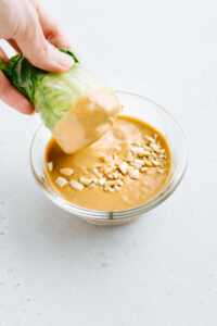 A small glass bowl on a grey backdrop with creamy peanut sauce garnished with peanuts and a hand dipping a summer roll in the sauce