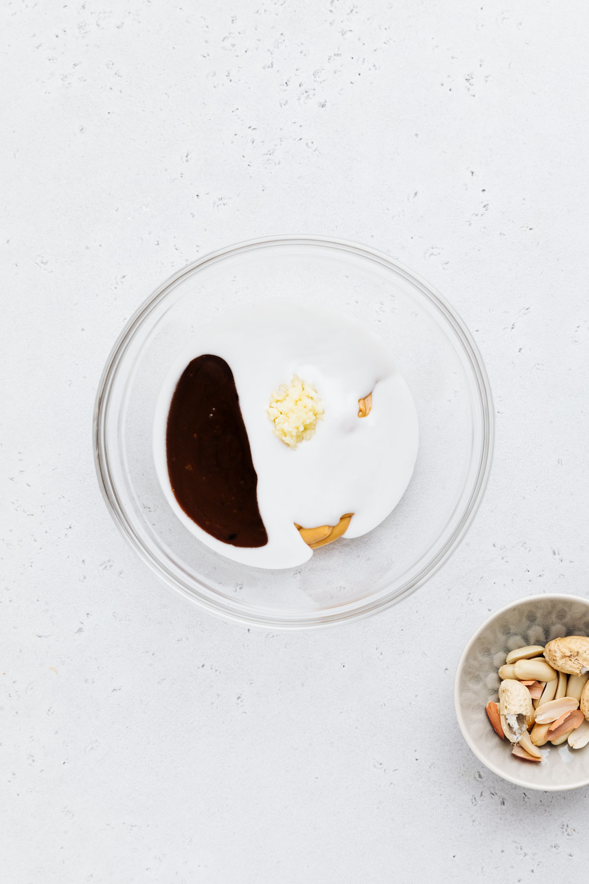 A glass bowl on a grey backdrop with creamy peanut butter, coconut milk, garlic and hoisin sauce in it and a small bowl with peanuts next to it