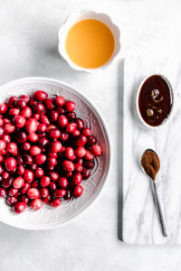 cranberry sauce ingredients with cranberries in a white bowl with a glass of orange juice and ingredients on a white marble cutting board