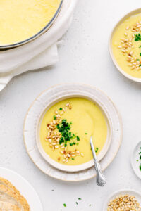 A bowl with vegan potato leek soup topped with roasted pine nuts and chives with a silver spoon in it and a pot with more soup shown in the corner