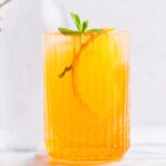 A glass with yellow Crodino Spritz with ice cubes garnished with mint and a slice of orange on a white marble backdrop.