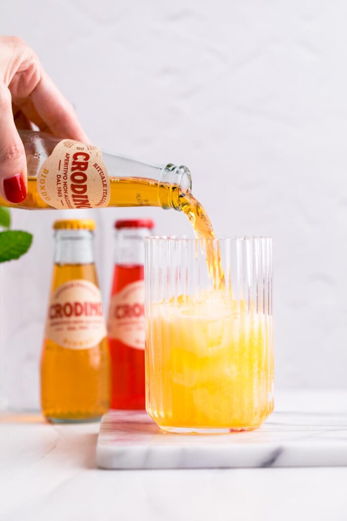 A hand pouring yellow Crodino into a glass with ice cubes on a white marble backdrop.