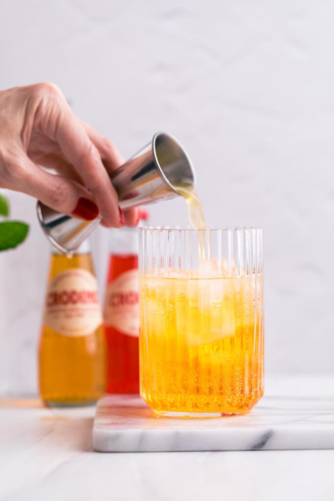 A hand pouring orange juice into a glass with ice cubes on a white marble backdrop.