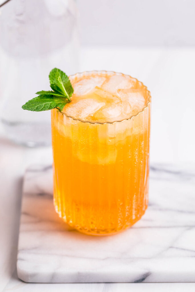 A glass with yellow Crodino Spritz with ice cubes garnished with mint on a white marble backdrop.