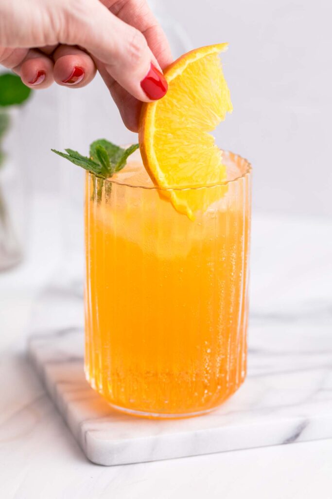 A glass with yellow Crodino Spritz with ice cubes garnished with mint and an orange slice being placed in the glass.