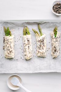 A row with four bundles with Asparagus with Puff Pastry on a baking tray with white parchment paper with a pastry brush and a small bowl next to it on a white backdrop.