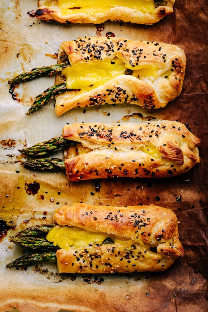 A row of three golden brown baked bundles Asparagus with Puff Pastry on a baking tray with browned parchment paper