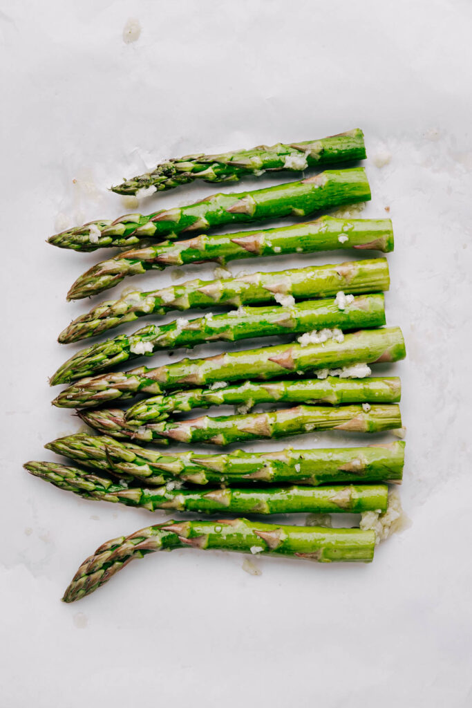 Asparagus in a row on white parchment paper with olive oil, lemon juice and minced garlic on the asparagus