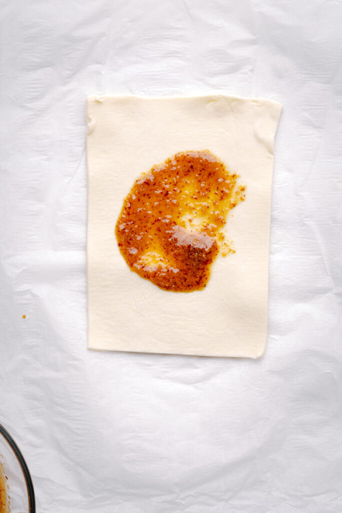 A square of puff pastry with a layer of mustard and maple syrup in the middle on white parchment paper