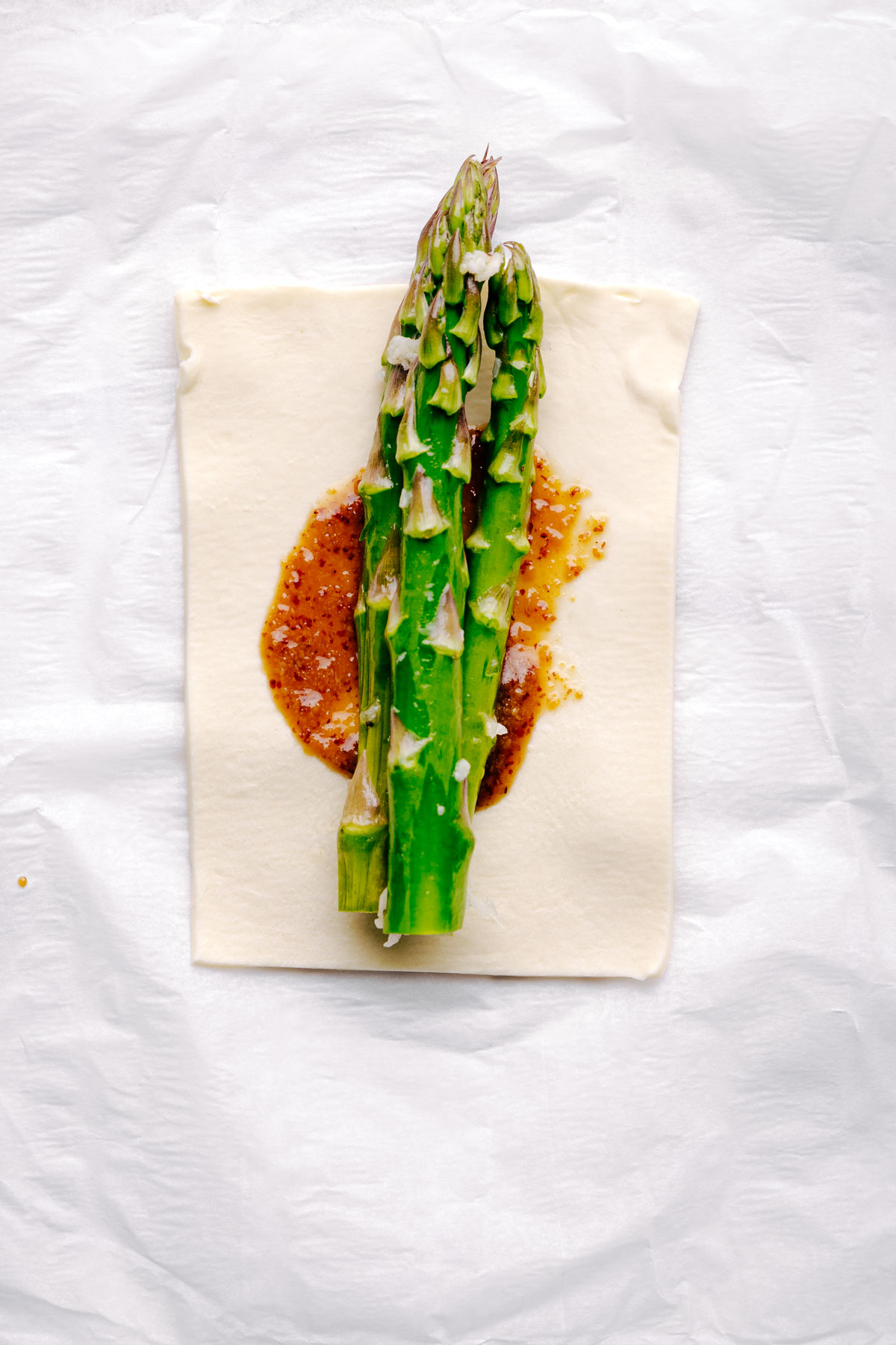 A square of puff pastry with a layer of mustard and maple syrup and three asparagus in the middle on white parchment paper