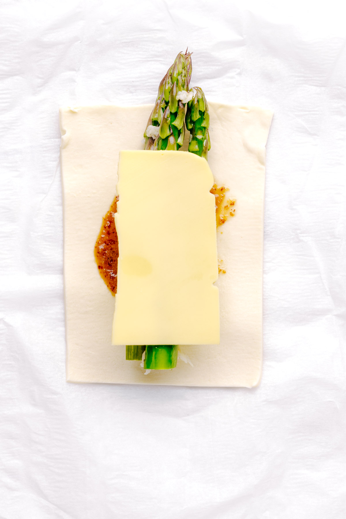 A square of puff pastry with a layer of mustard and maple syrup, three asparagus, and half a cheese slice in the middle on white parchment paper